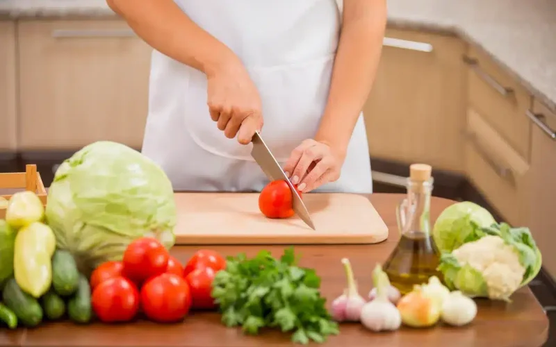 Person wearing a white apron slicing a tomato on a cutting board, surrounded by various fresh vegetables and a bottle of olive oil on a kitchen counter.