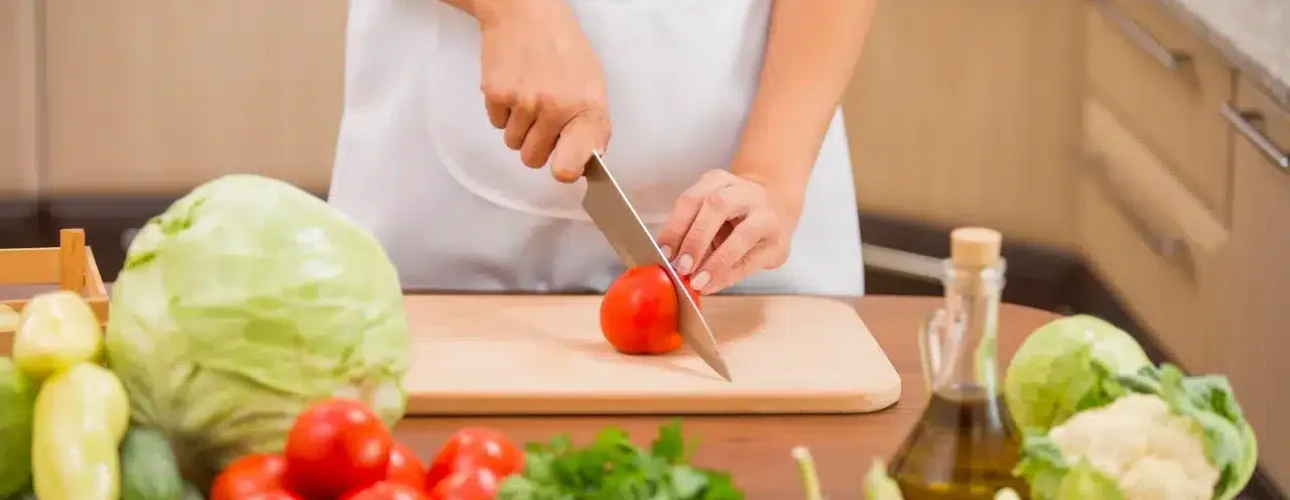 Person wearing a white apron slicing a tomato on a cutting board, surrounded by various fresh vegetables and a bottle of olive oil on a kitchen counter.