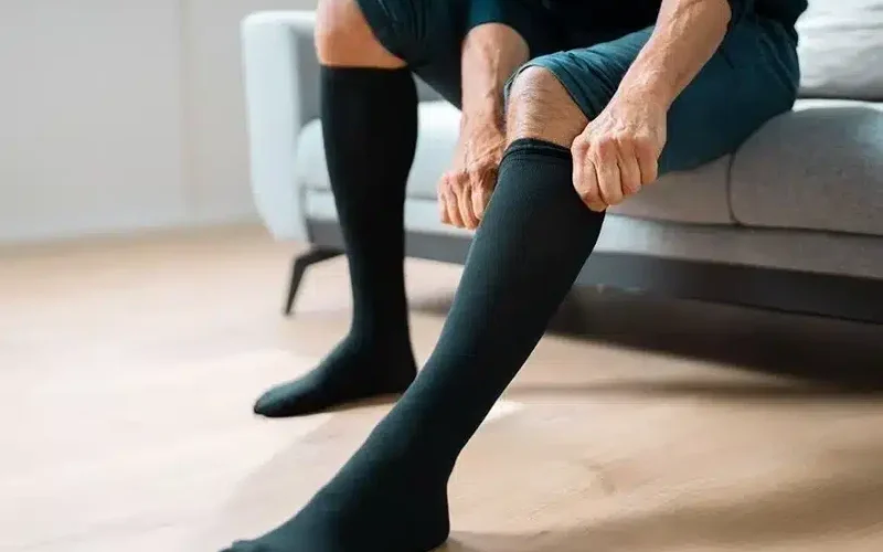 A person sitting on a couch pulls up black compression socks on their legs, with their feet resting on a wooden floor.
