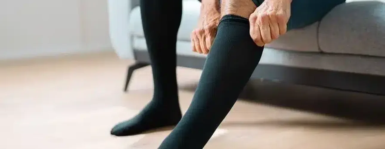 A person sitting on a couch pulls up black compression socks on their legs, with their feet resting on a wooden floor.
