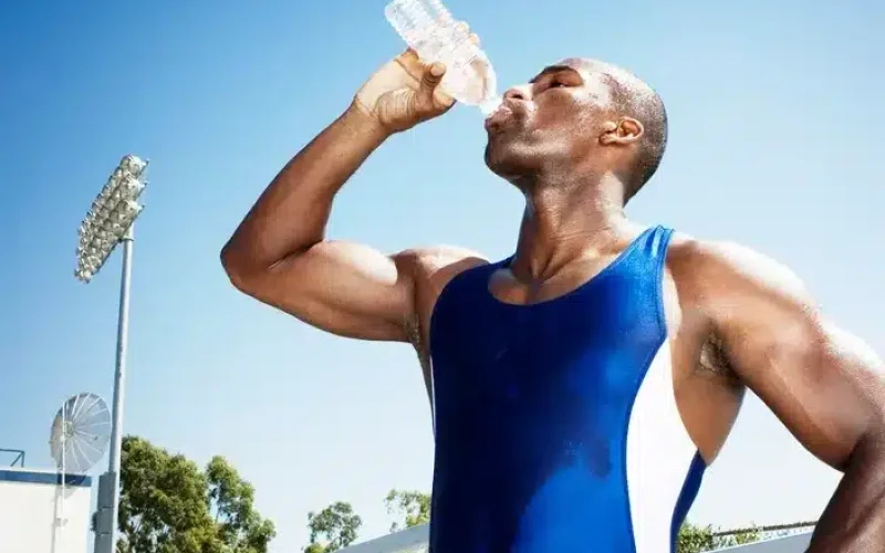 A person in athletic attire drinks water from a plastic bottle outdoors on a sunny day, with trees and stadium lights visible in the background.