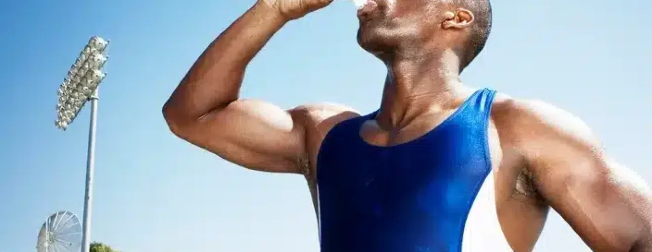 A person in athletic attire drinks water from a plastic bottle outdoors on a sunny day, with trees and stadium lights visible in the background.