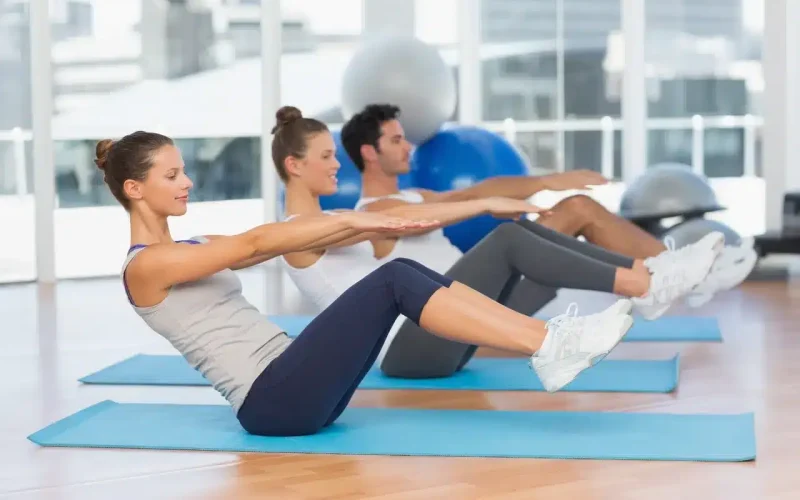 Three people exercise on yoga mats indoors, seated with legs raised and arms extended forward, practicing a balance or core workout.