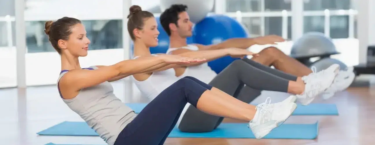 Three people exercise on yoga mats indoors, seated with legs raised and arms extended forward, practicing a balance or core workout.