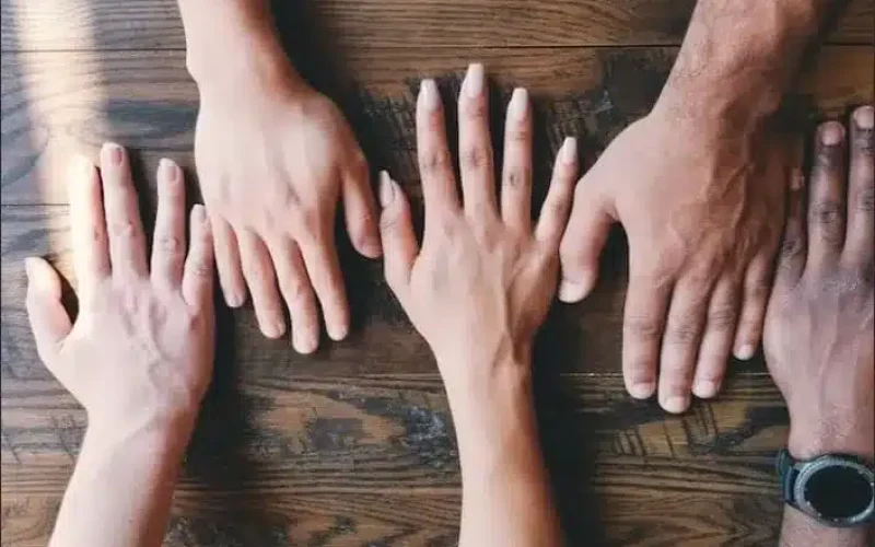 Four hands of different skin tones are placed flat on a wooden table, with a wristwatch visible on one wrist.