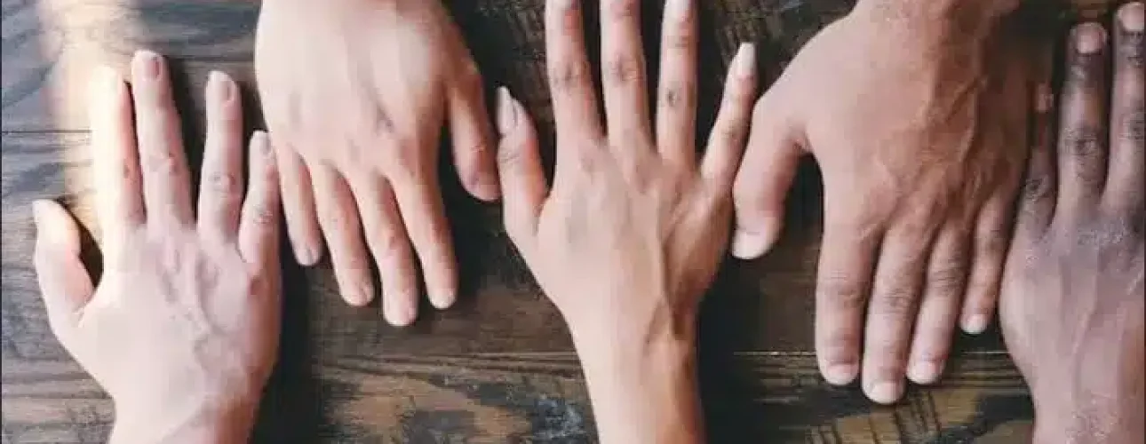Four hands of different skin tones are placed flat on a wooden table, with a wristwatch visible on one wrist.