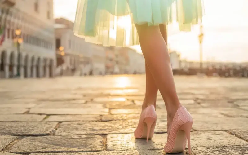 A person wearing a pastel tulle skirt and pink high heels walks on a stone-paved street at sunset.