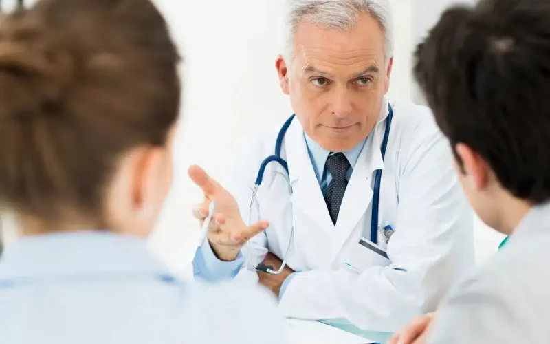 A male doctor in a white coat and stethoscope talks to two people seated across from him in an office setting.