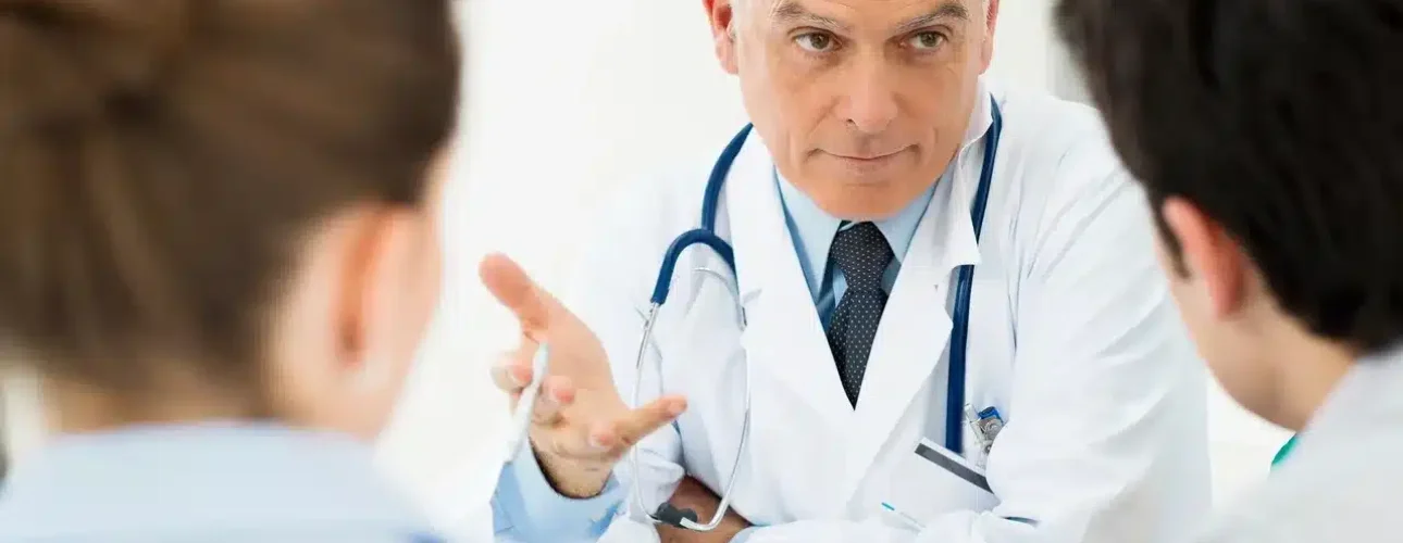 A male doctor in a white coat and stethoscope talks to two people seated across from him in an office setting.