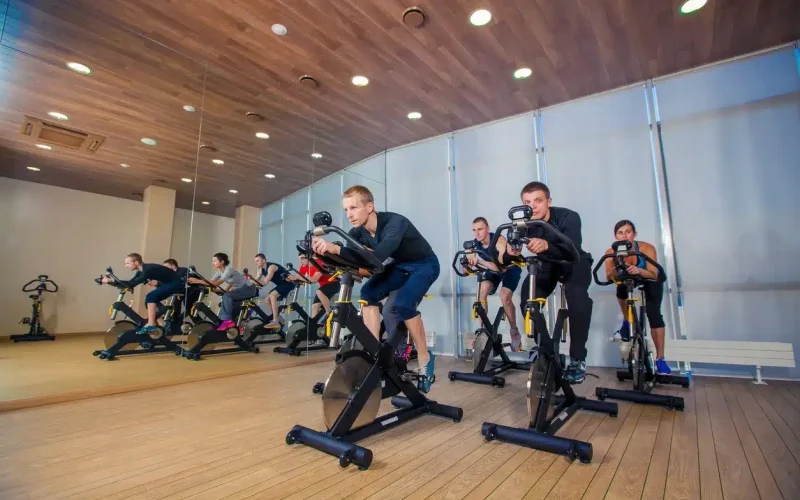 A group of people participate in a spin class, riding stationary bikes in a mirrored fitness studio with wood flooring and ceiling.