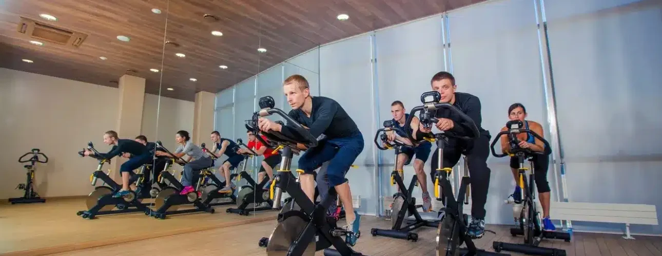 A group of people participate in a spin class, riding stationary bikes in a mirrored fitness studio with wood flooring and ceiling.