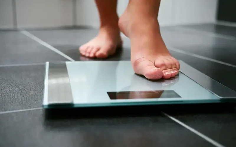 Close-up of a person stepping onto a glass digital bathroom scale on a dark tiled floor.