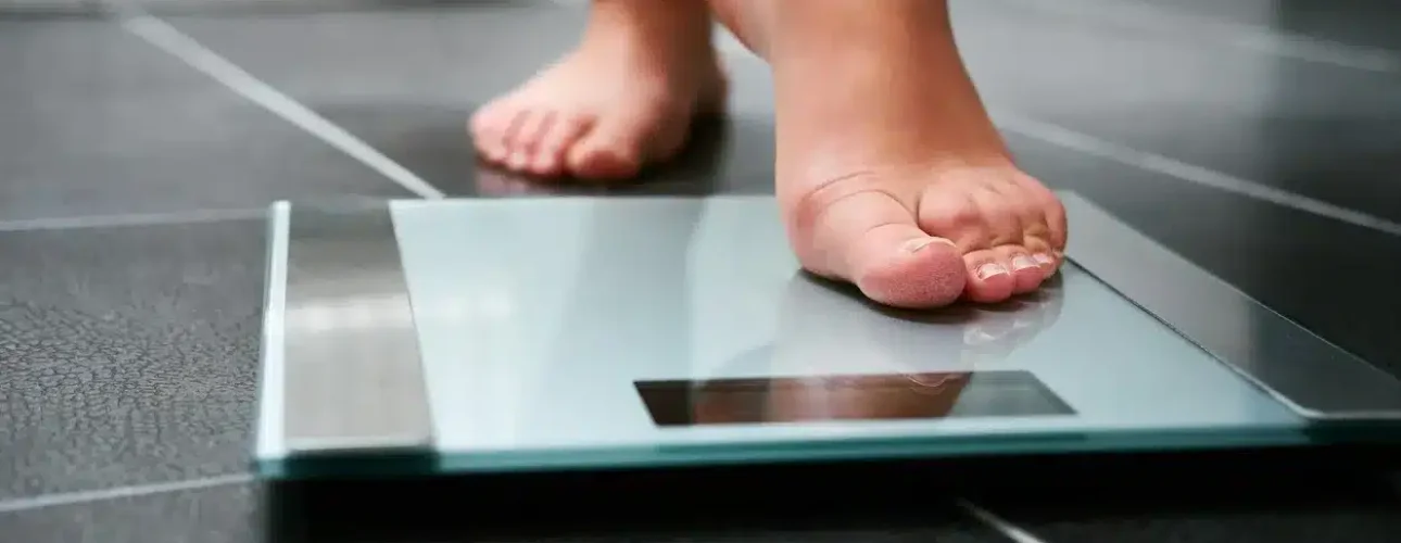Close-up of a person stepping onto a glass digital bathroom scale on a dark tiled floor.