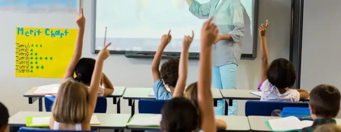 A teacher stands at the front of a classroom pointing at a whiteboard while several young students raise their hands.