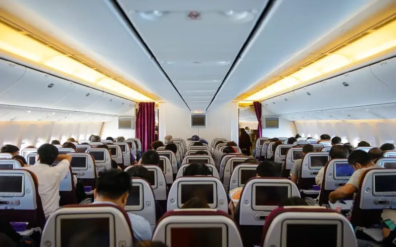 Interior of a commercial airplane showing passengers seated in economy class, with seat-back screens and overhead lighting visible.