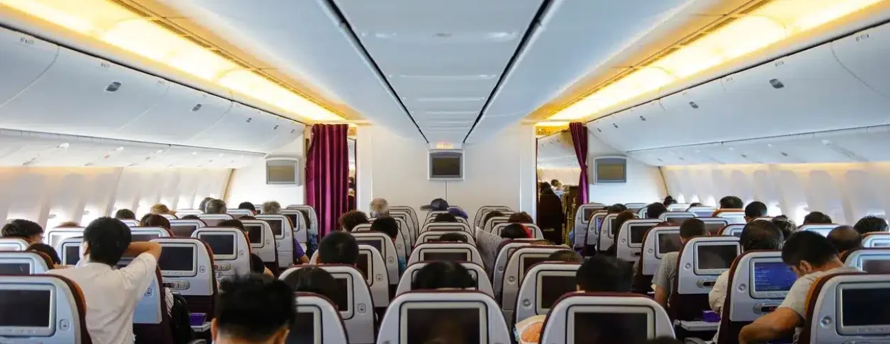 Interior of a commercial airplane showing passengers seated in economy class, with seat-back screens and overhead lighting visible.