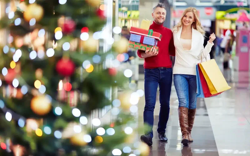A smiling couple walks through a shopping mall with shopping bags and gift boxes, passing a decorated Christmas tree.