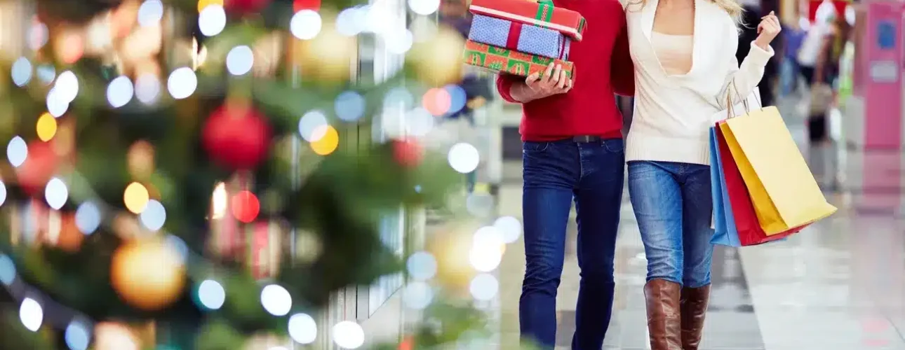 A smiling couple walks through a shopping mall with shopping bags and gift boxes, passing a decorated Christmas tree.