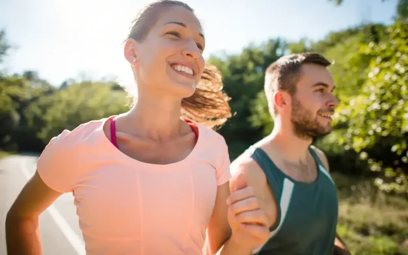 A woman and a man jog outdoors on a sunny day, smiling as they run along a path surrounded by greenery.