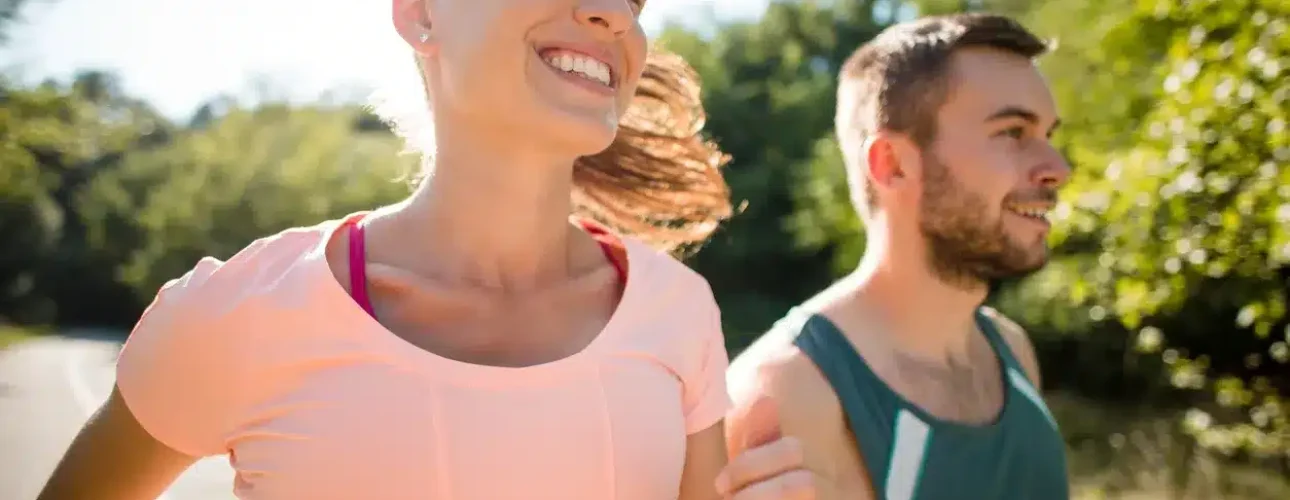 A woman and a man jog outdoors on a sunny day, smiling as they run along a path surrounded by greenery.