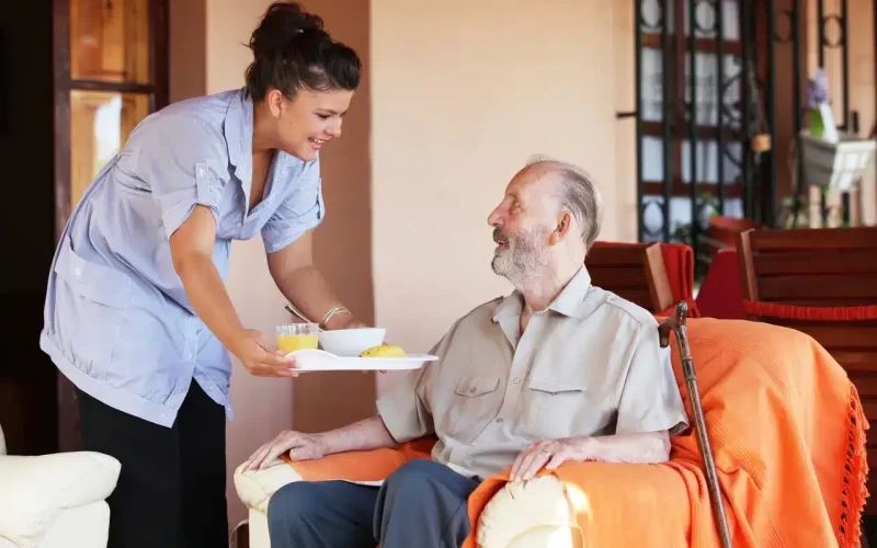 A caregiver is smiling while serving a meal on a tray to an elderly man sitting in an armchair with a cane beside him.