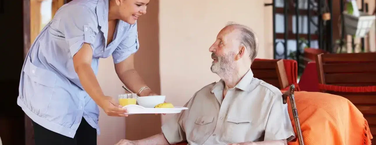 A caregiver is smiling while serving a meal on a tray to an elderly man sitting in an armchair with a cane beside him.