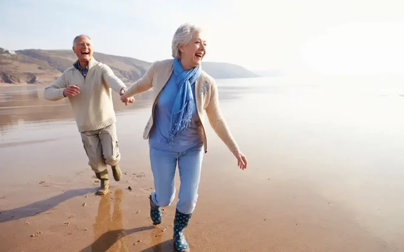 Two older adults smiling and holding hands while walking along a sandy beach, both wearing casual clothing and boots.