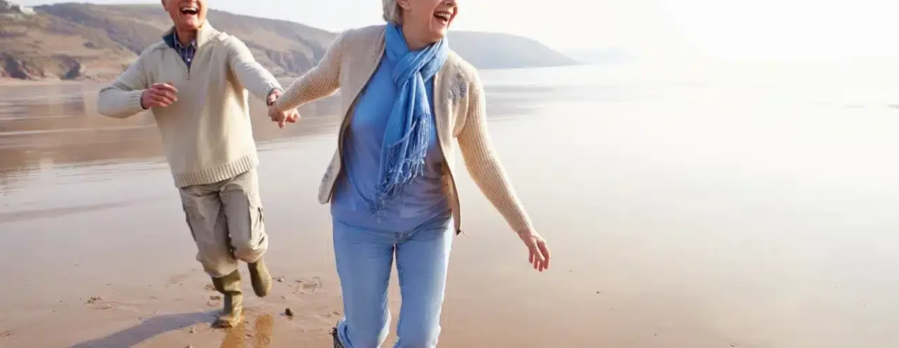 Two older adults smiling and holding hands while walking along a sandy beach, both wearing casual clothing and boots.