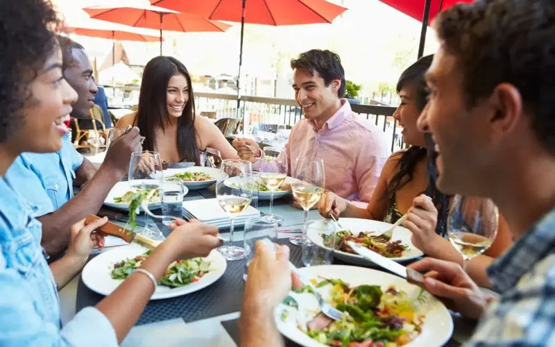 A group of six people sit at an outdoor restaurant table, eating salads and meals, drinking wine, and conversing under red umbrellas.