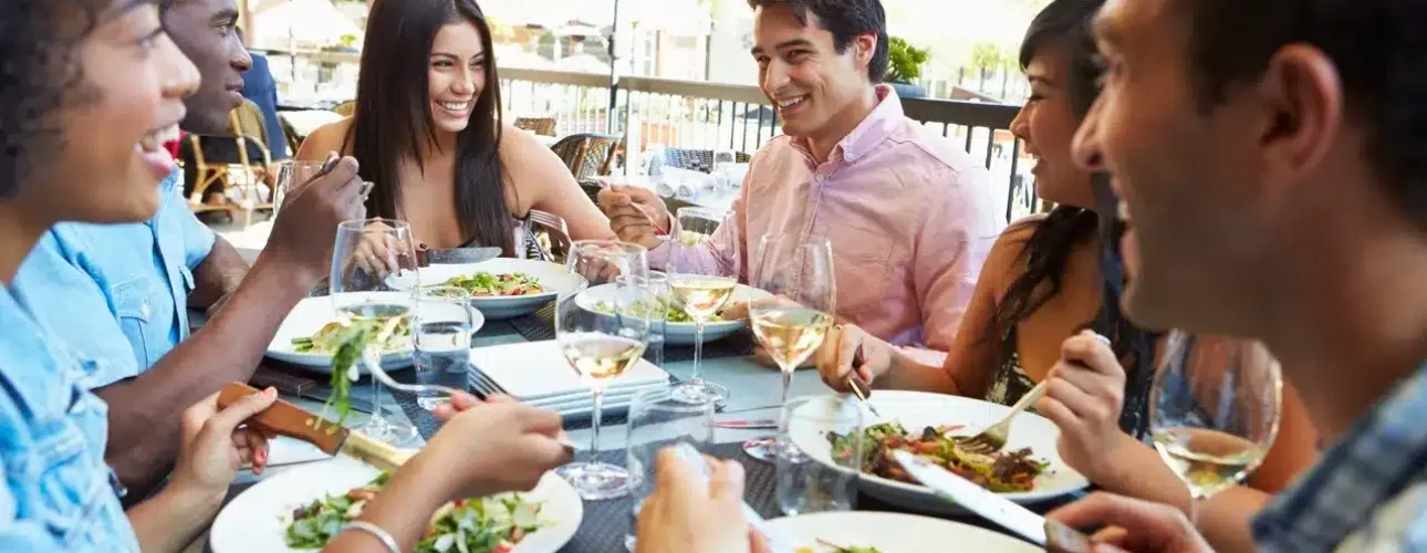 A group of six people sit at an outdoor restaurant table, eating salads and meals, drinking wine, and conversing under red umbrellas.