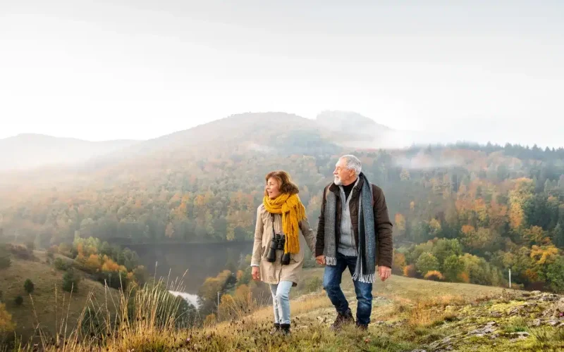 An older couple walks together on a grassy hillside with autumn trees and misty mountains in the background. The woman wears a yellow scarf and binoculars around her neck.
