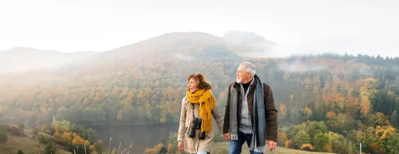 An older couple walks together on a grassy hillside with autumn trees and misty mountains in the background. The woman wears a yellow scarf and binoculars around her neck.