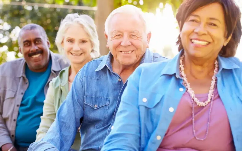 Four older adults, two men and two women, sit outdoors in a row, smiling at the camera with green trees in the background.