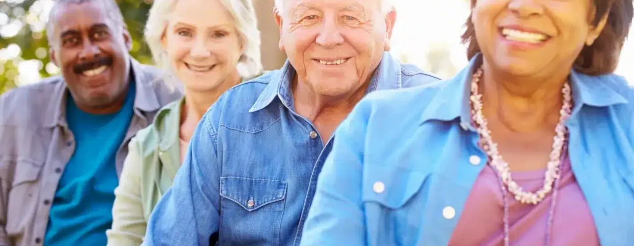 Four older adults, two men and two women, sit outdoors in a row, smiling at the camera with green trees in the background.