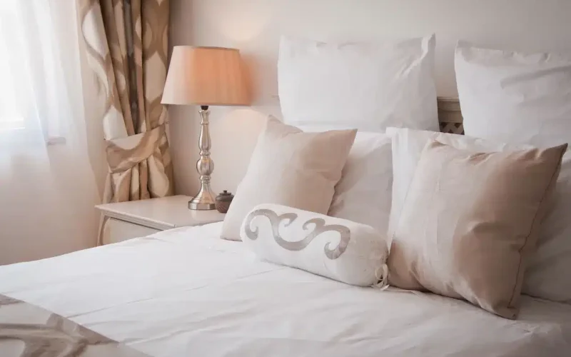 A neatly made bed with white and beige pillows, a decorative cushion, and a bedside table with a lamp in a softly lit bedroom.