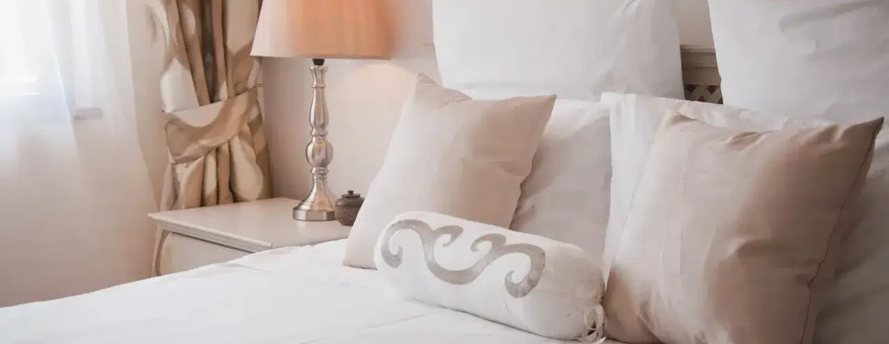 A neatly made bed with white and beige pillows, a decorative cushion, and a bedside table with a lamp in a softly lit bedroom.