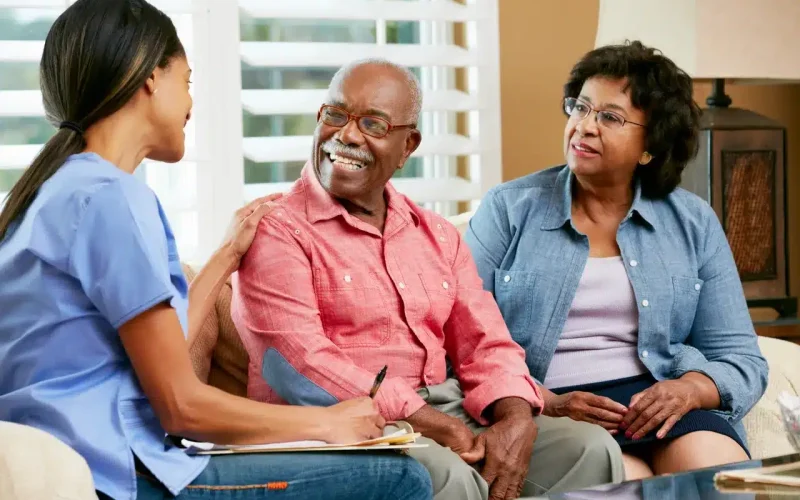 A nurse speaks with an elderly man and woman sitting on a couch, smiling and holding a clipboard in a well-lit living room.