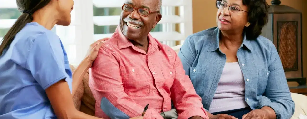 A nurse speaks with an elderly man and woman sitting on a couch, smiling and holding a clipboard in a well-lit living room.