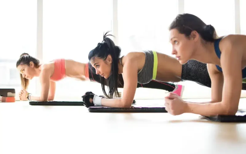 Three women are exercising indoors on yoga mats, performing planks and push-ups in a bright room with large windows.