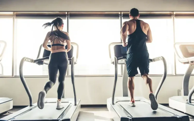 A woman and a man run side by side on treadmills in a gym with large windows letting in natural light.
