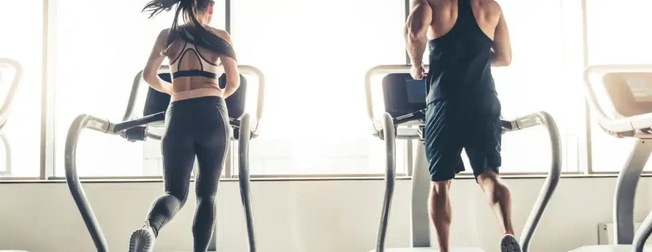 A woman and a man run side by side on treadmills in a gym with large windows letting in natural light.