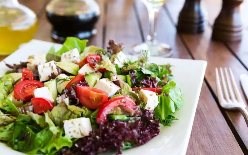 A plate of fresh salad with mixed greens, cherry tomatoes, cucumber, and cubes of cheese on a wooden table, with utensils and glassware in the background.