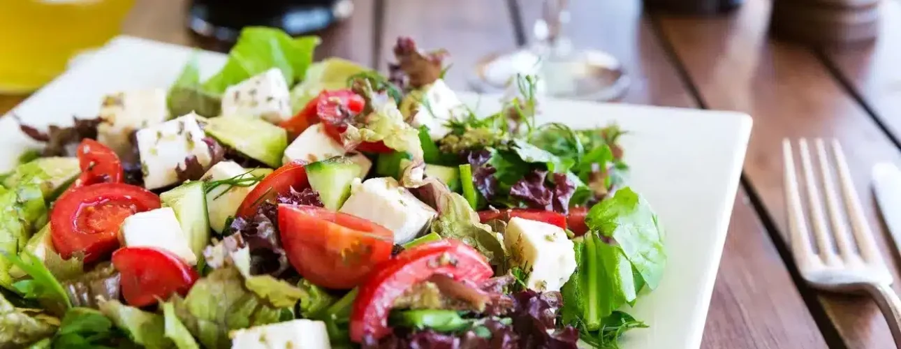 A plate of fresh salad with mixed greens, cherry tomatoes, cucumber, and cubes of cheese on a wooden table, with utensils and glassware in the background.