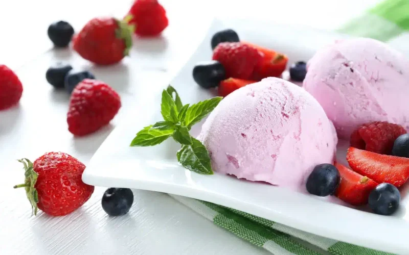 Two scoops of pink ice cream on a white plate, garnished with fresh strawberries, blueberries, raspberries, and a sprig of mint.