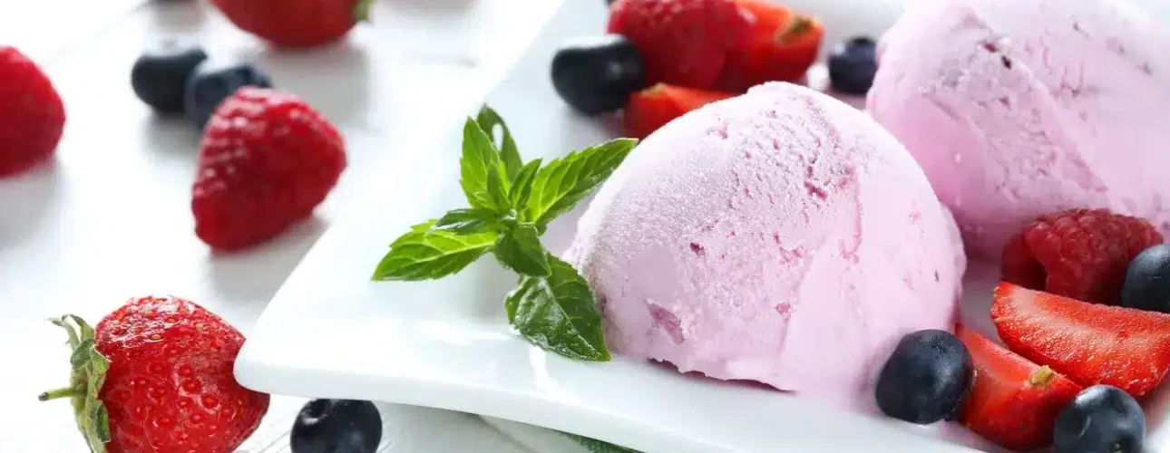 Two scoops of pink ice cream on a white plate, garnished with fresh strawberries, blueberries, raspberries, and a sprig of mint.