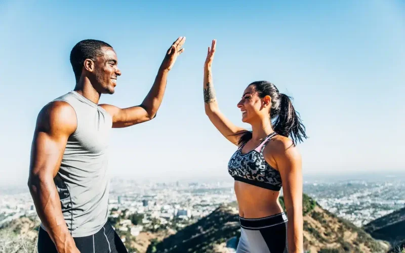 Two people in athletic wear give each other a high five outdoors, with hills and a city skyline in the background under a clear blue sky.