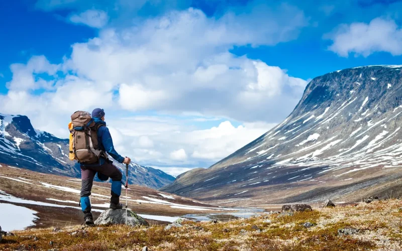 A hiker with a backpack and trekking poles stands on rocky terrain, looking at a snow-capped mountain under a partly cloudy blue sky.