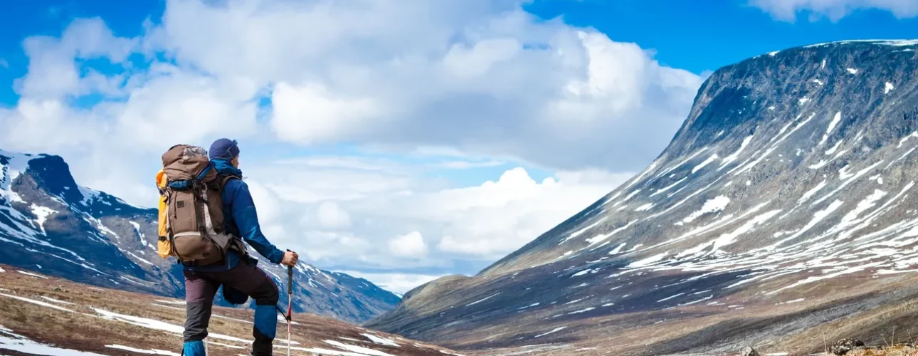A hiker with a backpack and trekking poles stands on rocky terrain, looking at a snow-capped mountain under a partly cloudy blue sky.