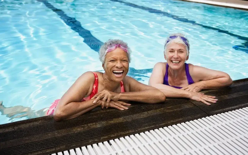 Two older women wearing swimsuits and swim goggles smile and lean on the edge of an indoor swimming pool.
