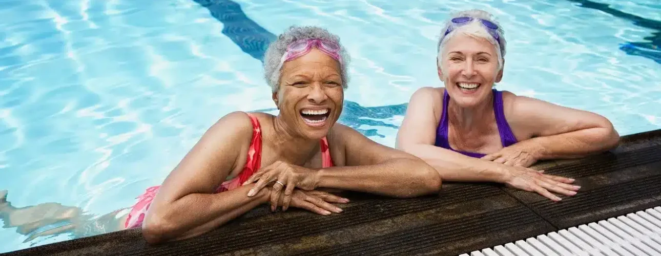 Two older women wearing swimsuits and swim goggles smile and lean on the edge of an indoor swimming pool.